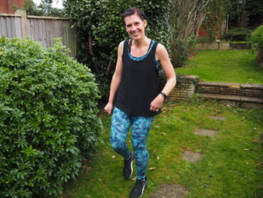 Smiling dark haired girl in graden next to a bush, wearing tropical blue leggings and black vest top