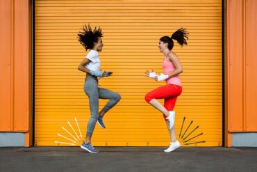 Two ladies in their early thirties dressed in fitness gear high knee running on the spot. Yello lined background, one on right wearing 3/4 length red leggings, pink vest top. One on left wearing full length mid grey leggins and a white t-shirt. Trying to increase their metabolism