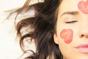 Close up of half a young brunette lady's face. her eye is closed and her hair is spread out on a white sheet. She has a rose petal that looks like a sliced strawberry on her cheek and forehead.