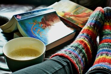 close up of a womans feet in brightly coloured striped woolen socks on a bed with two open books and a cup of coffee next to her.