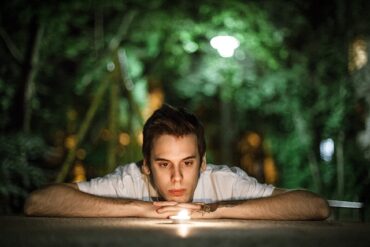 a young man resting his arms on a table looking into the flame of a candle. Going through the process of healing and learning he is not at fault.