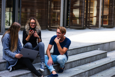 group of three college students sat on outsde steps at fron tof college building. They are laughing together and looking as though they are stress free